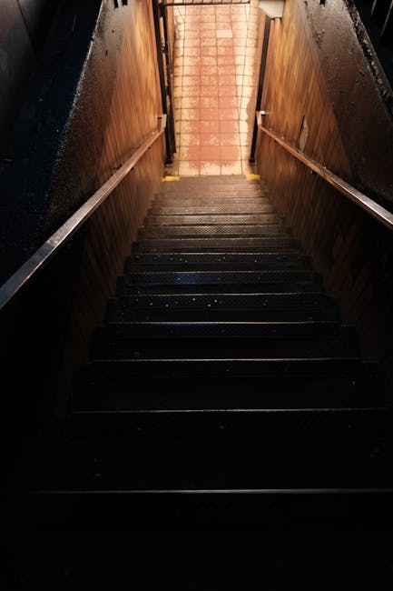 A narrow staircase with dark, non-slip wooden steps ascending towards a dimly-lit area, with wooden paneling along the walls and metal handrails on each side. The lighting emphasizes the texture of the steps and the wood, highlighting the cleanliness and maintenance. This staircase, part of a residential or commercial building, presents access challenges due to its tight space and narrow width, which cleanersw9.co.uk manages through careful surface cleaning and safety considerations, ensuring an accessible and hygienic environment. The overall scene underscores the importance of effective cleaning to maintain safety and appearance in confined stairwells within Maida Vale, W9, in line with domestic cleaning and surface sanitisation practices.