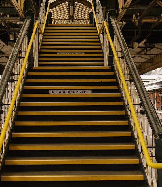The image displays a narrow, spiraling metal staircase with worn blue-painted steps that are peeling, revealing the underlying material. The staircase is set within a small enclosed space with white walls, one of which shows signs of wear and patches of peeling paint. Adjacent to the staircase, a vertical white pipe runs alongside the wall. The area is lit by natural light coming from an unseen source above, highlighting the somewhat aged and utilitarian appearance of the stairs. This scene illustrates the challenges of cleaning and maintaining tight, multi-level pathways in residential or commercial settings, aligning with the context of access problems for Maida Vale cleaners narrow stairs, W9, as managed by Cleaners W9, specialists in surface cleaning and hygiene solutions.