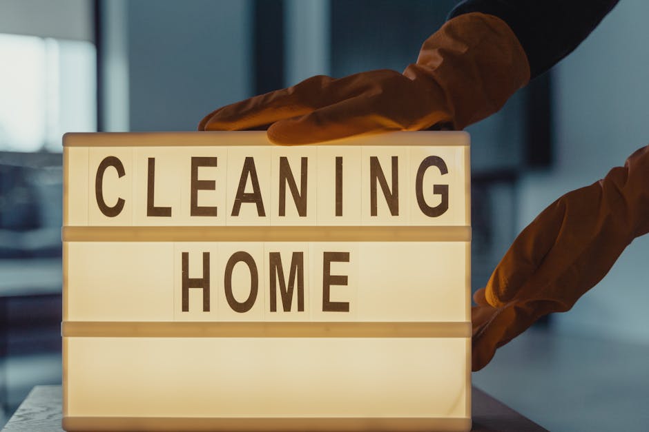 A person wearing brown cleaning gloves is adjusting a lit-up signboard displaying the words 'CLEANING HOME' in bold black letters. The signboard is positioned on a table in a room with a modern, minimalist background, featuring a dark blue wall and natural light coming through a window. The surface of the table appears smooth and clean, emphasizing quality surface cleaning and maintenance. The scene reflects professional domestic cleaning services provided by Cleaners W9, focusing on thorough sanitisation and deep cleaning, suitable for residential spaces such as kitchens or living rooms.