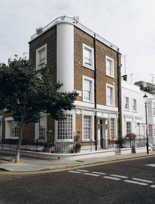 Exterior view of a multi-story residential building on Lauderdale Road in Maida Vale, W9, featuring a corner section with brick and white-painted surfaces, large windows with white frames, and a rooftop terrace with a metal railing. The building's ground level includes a white entrance door, potted plants, and black metal fencing along the sidewalk. The street in front is paved with asphalt and marked with pedestrian crossings, while a tree with dark green foliage partially obscures the left side of the building. The overall scene showcases a well-maintained, clean, and tidy facade typical of the Maida Vale area, emphasizing the importance of building exterior maintenance and cleanliness, as highlighted by professional cleaning services such as those provided by Cleaners W9.