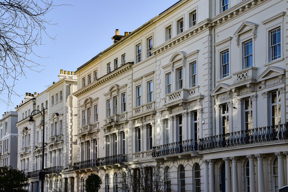 Exterior view of a row of Victorian-style white residential buildings in Maida Vale, W9, featuring ornate window frames, decorative cornices, black wrought iron balconies, and tall sash windows. The buildings are set against a clear blue sky, with leafless trees and traditional black street lamps visible in the foreground. The clean, well-maintained façade reflects recent surface cleaning and restoration, typical of professional domestic and commercial cleaning projects by Cleaners W9. The image emphasizes the building's historic architecture and pristine condition, consistent with high-quality cleaning and maintenance standards for properties in the Lauderdale Road area.