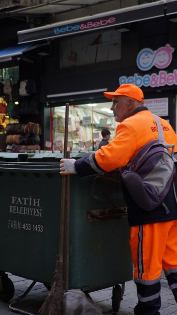 A sanitation worker dressed in an orange high-visibility uniform with reflective stripes and a matching orange cap is tending to a large green waste bin on a city street. The worker is holding a wooden broom and appears to be collecting waste or debris. The background features a storefront with signage reading 'bebe & Bebe' in pink and blue lettering, along with display windows and posters. The scene is set during the daytime, with natural light illuminating the area, highlighting the cleanliness and organization of the street environment. The worker's attire and the waste management activity reflect a focus on hygiene and urban sanitation, aligning with the topic of licensing and waste compliance for cleaners in Maida Vale, W9, as emphasized by Cleaners W9.