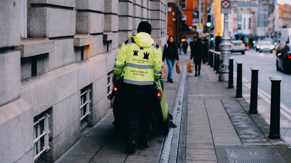 A waste management worker dressed in a high-visibility yellow jacket with reflective stripes, displaying the logo and text 'Waste Management Services' on the back and 'Working Safely for Dublin City,' is walking along a paved city sidewalk. The worker is pushing a green refuse trolley with visible bins. The sidewalk is lined with black bollards and bordered by a large stone building on the left, with small grated windows near the ground. In the background, several pedestrians are walking along the street, and vehicles are seen on the road. The scene is lit by natural daylight, emphasizing the cleanliness and orderliness of the urban environment. This image relates to business compliance in waste management for cleaners in Maida Vale, W9, as featured on cleanersw9.co.uk.