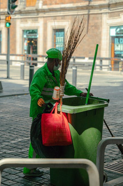 A sanitation worker dressed in an orange high-visibility uniform with reflective stripes and a matching orange cap is tending to a large green waste bin on a city street. The worker is holding a wooden broom and appears to be collecting waste or debris. The background features a storefront with signage reading 'bebe & Bebe' in pink and blue lettering, along with display windows and posters. The scene is set during the daytime, with natural light illuminating the area, highlighting the cleanliness and organization of the street environment. The worker's attire and the waste management activity reflect a focus on hygiene and urban sanitation, aligning with the topic of licensing and waste compliance for cleaners in Maida Vale, W9, as emphasized by Cleaners W9.