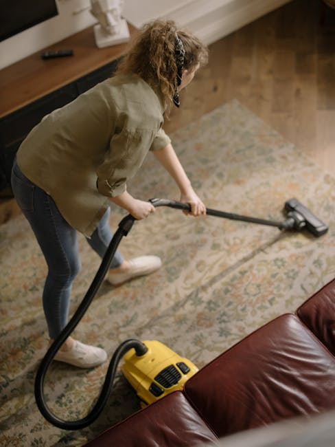 A woman operating a yellow vacuum cleaner with a black hose and floor attachment, cleaning a patterned area rug on a wooden floor in a living room. She is dressed in casual clothing, including a beige jacket, dark jeans, and white sneakers. The room features a dark wooden sideboard, a white decorative sculpture, and natural lighting. The scene illustrates domestic surface cleaning as part of professional services by Cleaners W9, showcasing deep cleaning of carpets and rugs to ensure cleanliness and hygiene in residential spaces.