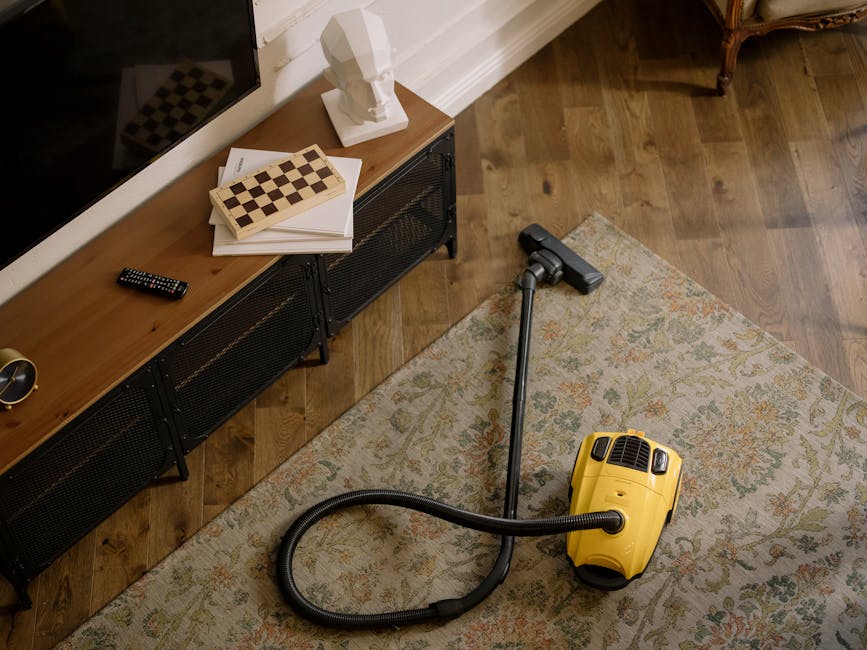 A yellow vacuum cleaner with a black hose and metal wand attachment is positioned on a patterned area rug in a living room, next to a wooden sideboard with black metal legs. On top of the sideboard, there is a white sculptural bust, some stacked papers, a chessboard, and a TV remote control. The room features wooden flooring and a white baseboard along the wall, with soft natural lighting illuminating the space, demonstrating surface cleaning and maintenance by Cleaners W9 in Maida Vale, W9.
