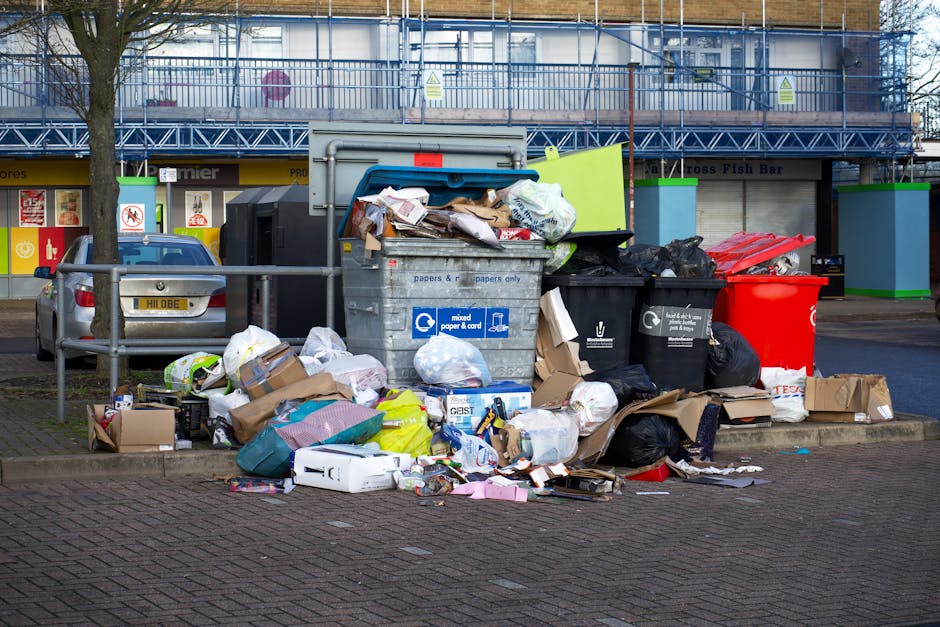 A large pile of mixed waste and bulky cardboard boxes overflowing from a grey recycling bin labeled for papers and cardboard, situated on a paved urban sidewalk with a metal railing. Surrounding the bin are additional black and red waste bags, scattered packaging materials, and cardboard boxes, indicating a cluttered area. In the background, a parked silver car is visible alongside a commercial building with storefronts, a tree to the left, and a construction scaffolding structure above with safety signs. The scene appears unclean with debris and waste materials partly strewn on the pavement, emphasizing the importance of proper waste management in residential or commercial cleaning contexts. The image highlights the need for adhering to waste disposal regulations, as detailed in the Maida Vale W9 bulky waste rules for cleaning companies, with professional services like those offered by Cleaners W9 ensuring hygienic cleanup and waste removal.