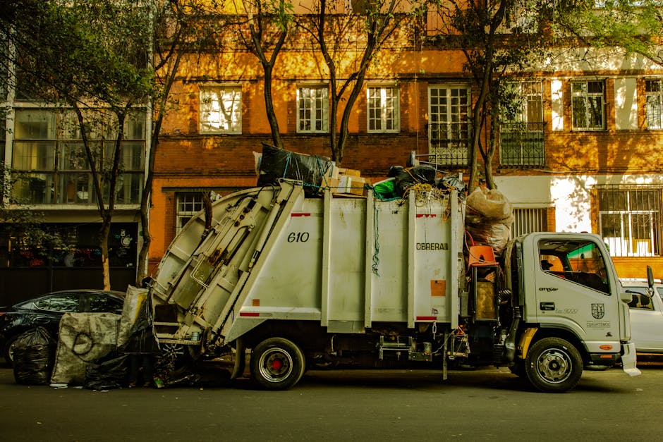 A white waste collection truck parked on a residential street during daytime, with an orange brick building and several trees casting shadows in the background. The truck is loaded with bulky waste items such as large bags, cardboard boxes, and miscellaneous rubbish, with some waste placed on the ground nearby. The truck's side displays the company name 'Dorebal' and service identification numbers. The scene is well-lit with natural sunlight, highlighting the clean surfaces of the truck and the scattered waste, representative of domestic or commercial waste collection practices in accordance with Maida Vale W9 bulky waste rules for cleaning companies, as promoted by Cleaners W9.