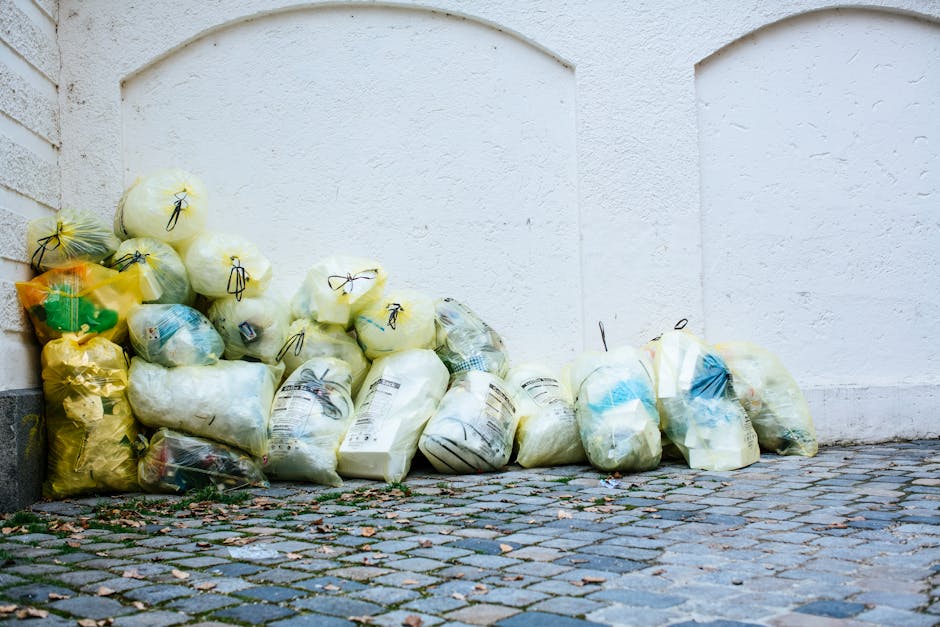 A large pile of mixed waste and bulky cardboard boxes overflowing from a grey recycling bin labeled for papers and cardboard, situated on a paved urban sidewalk with a metal railing. Surrounding the bin are additional black and red waste bags, scattered packaging materials, and cardboard boxes, indicating a cluttered area. In the background, a parked silver car is visible alongside a commercial building with storefronts, a tree to the left, and a construction scaffolding structure above with safety signs. The scene appears unclean with debris and waste materials partly strewn on the pavement, emphasizing the importance of proper waste management in residential or commercial cleaning contexts. The image highlights the need for adhering to waste disposal regulations, as detailed in the Maida Vale W9 bulky waste rules for cleaning companies, with professional services like those offered by Cleaners W9 ensuring hygienic cleanup and waste removal.