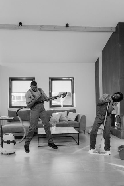 Two professional cleaners from Cleaners W9 are performing deep surface cleaning in a modern living room. The cleaner on the left is using a vacuum cleaner on the light-colored tile floor, which appears clean and well-maintained, with minimal dust or debris visible. They are wearing protective masks and uniform, emphasizing hygiene and safety. The other cleaner on the right is mopping the floor with a mop, focusing on a particular section near a built-in fireplace, which has a sleek, contemporary design with a metallic finish. The room features a neutral color palette with white walls, wooden accent panels, and a large window allowing natural light to brighten the space. The sofa in the background has plush cushions, and the overall environment looks tidy and freshly cleaned, illustrating professional domestic cleaning services that include surface sanitation and thorough maintenance. The scene portrays a professional, hygienic cleaning process aligned with surface and deep cleaning standards, reflecting the top-tier service offered by Cleaners W9 for residential spaces around Maida Vale W9. This image visually represents the core activities involved in thorough domestic cleaning, emphasizing hygiene, cleanliness, and the use of appropriate tools and materials.