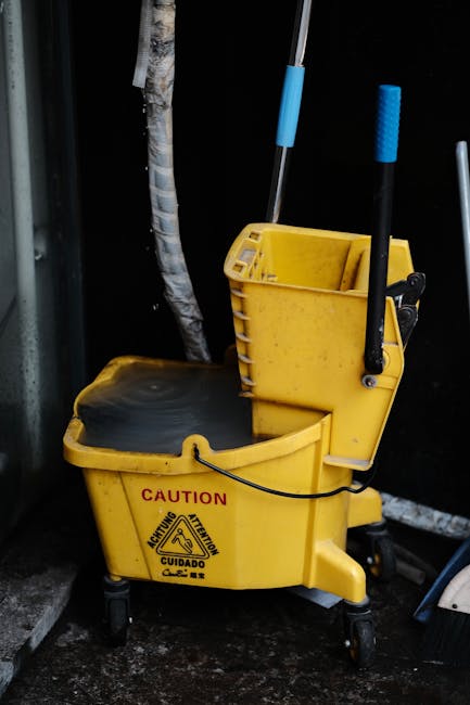 An image of a yellow commercial cleaning mop bucket with a black floor scrubber pad resting inside, positioned on a dark, wet surface outdoors. The bucket has a black handle and a caution warning label on the front, and it is equipped with a wringer and built-in storage compartments holding cleaning tools such as mops with blue handles. Nearby, there are partially visible black walls and a tree trunk, suggesting a commercial or industrial entrance area. The overall scene emphasizes surface cleaning and maintenance equipment used by Cleaners W9 for emergency cleaning and sanitisation services in Maida Vale, W9.