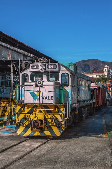 A metallic electric locomotive with the number 641 visible on the front, displaying a clean and polished surface with a light grey exterior accented by yellow and black safety markings on the front buffer beam. The train is situated on railway tracks adjacent to a platform, with the surrounding area featuring industrial and urban elements, including other freight cars attached behind the locomotive. The scene is set under bright daylight, providing clear visibility of the train's sleek surface and well-maintained appearance. Cleaners W9 specializes in surface cleaning and deep cleaning, ensuring hygiene and cleanliness for residential and commercial spaces, as seen reflected in their professional approach to maintaining high standards of sanitation.