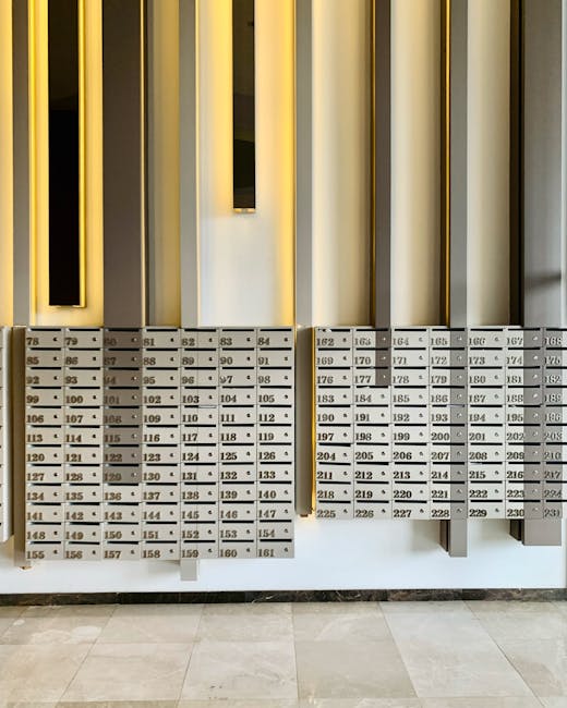 A row of metallic mailboxes with numbered compartments mounted on a white wall in a residential building hallway. The mailboxes have a modern design with small, rectangular opening handles. Above the mailboxes, there are vertical decorative panels in shades of black and gold, providing an elegant backdrop. The floor is tiled with light-colored, smooth ceramic tiles, and the area appears clean and well-maintained, reflecting the quality of surface cleaning and hygiene maintained by Cleaners W9 in preparation for the Sutherland Avenue end of tenancy cleaning in Maida Vale, W9.